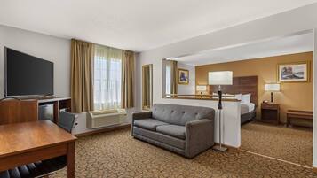 A hotel room with a flat-screen TV, a sofa, a desk with a chair, a bed, a bedside table with a lamp, and a patterned carpet.