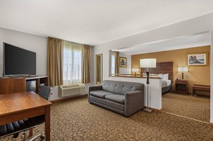 Habitación de hotel con televisor de pantalla plana, sofá, escritorio con silla, cama, mesita de noche con lámpara y alfombra estampada.