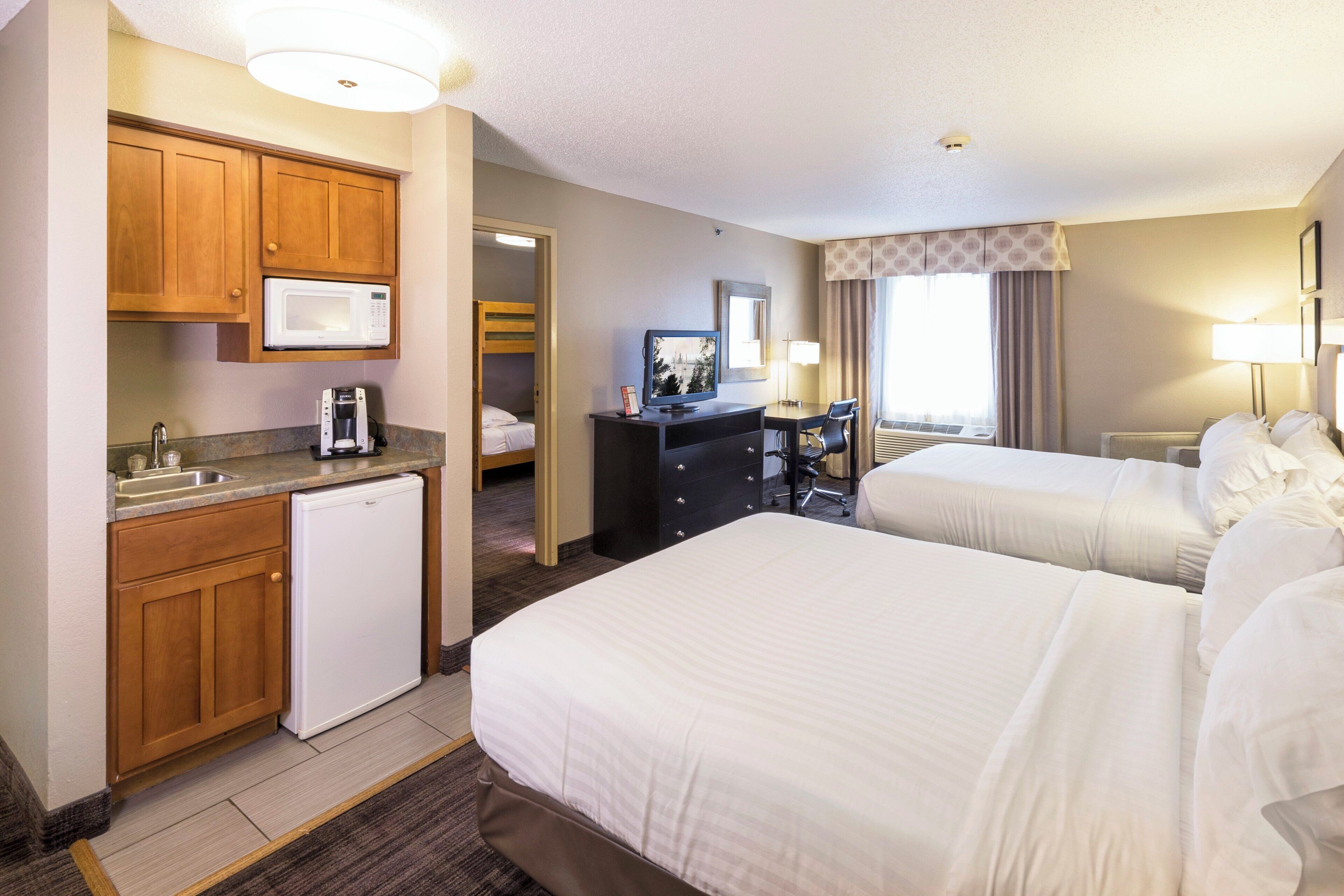 A hotel room with two beds, a kitchenette, a desk, and a television.
