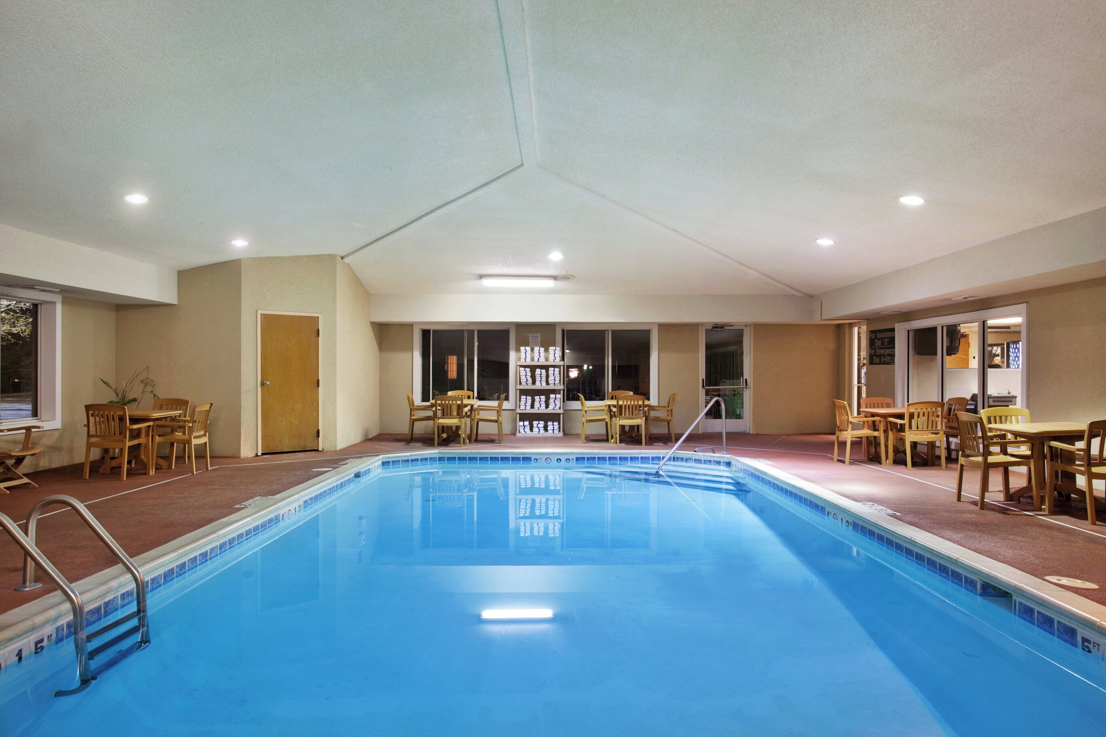 A hotel pool area with a clear blue pool, wooden chairs, and tables arranged around the pool.