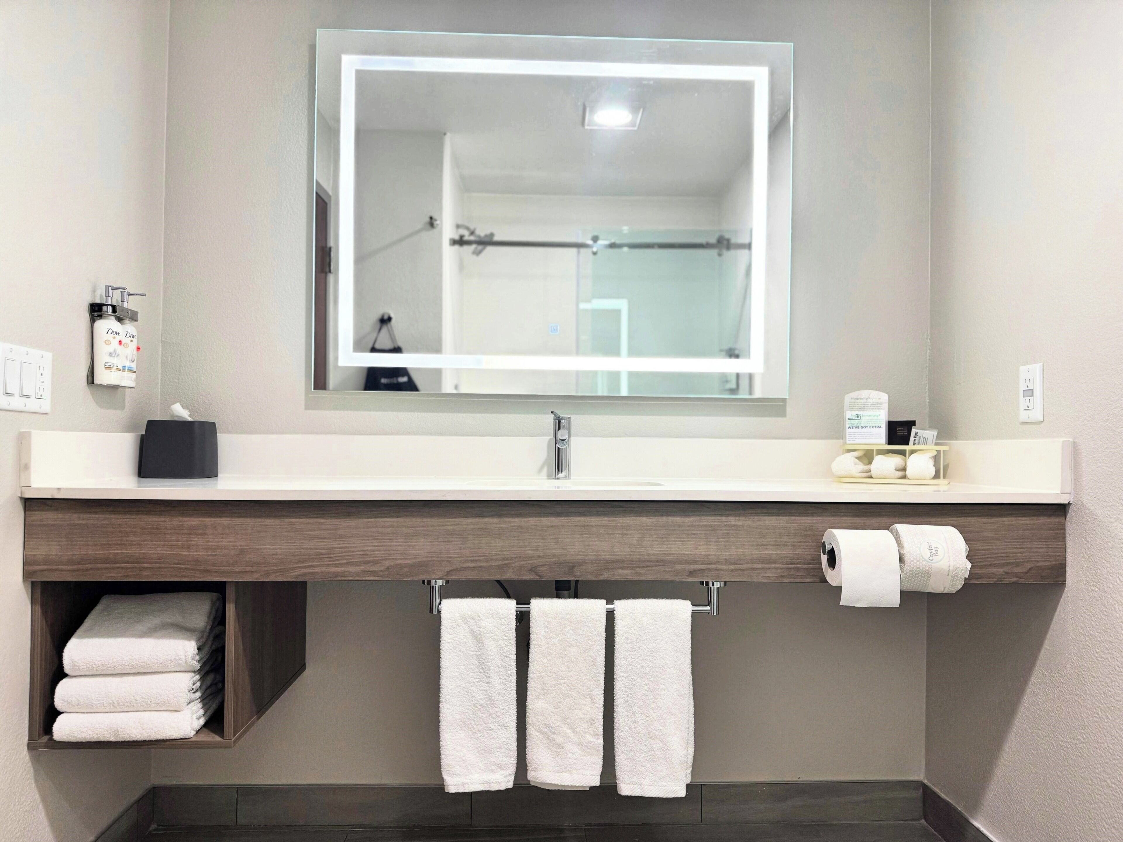 Une salle de bain moderne avec un grand miroir, un lavabo, un porte-serviettes et une étagère où sont pliés des serviettes.