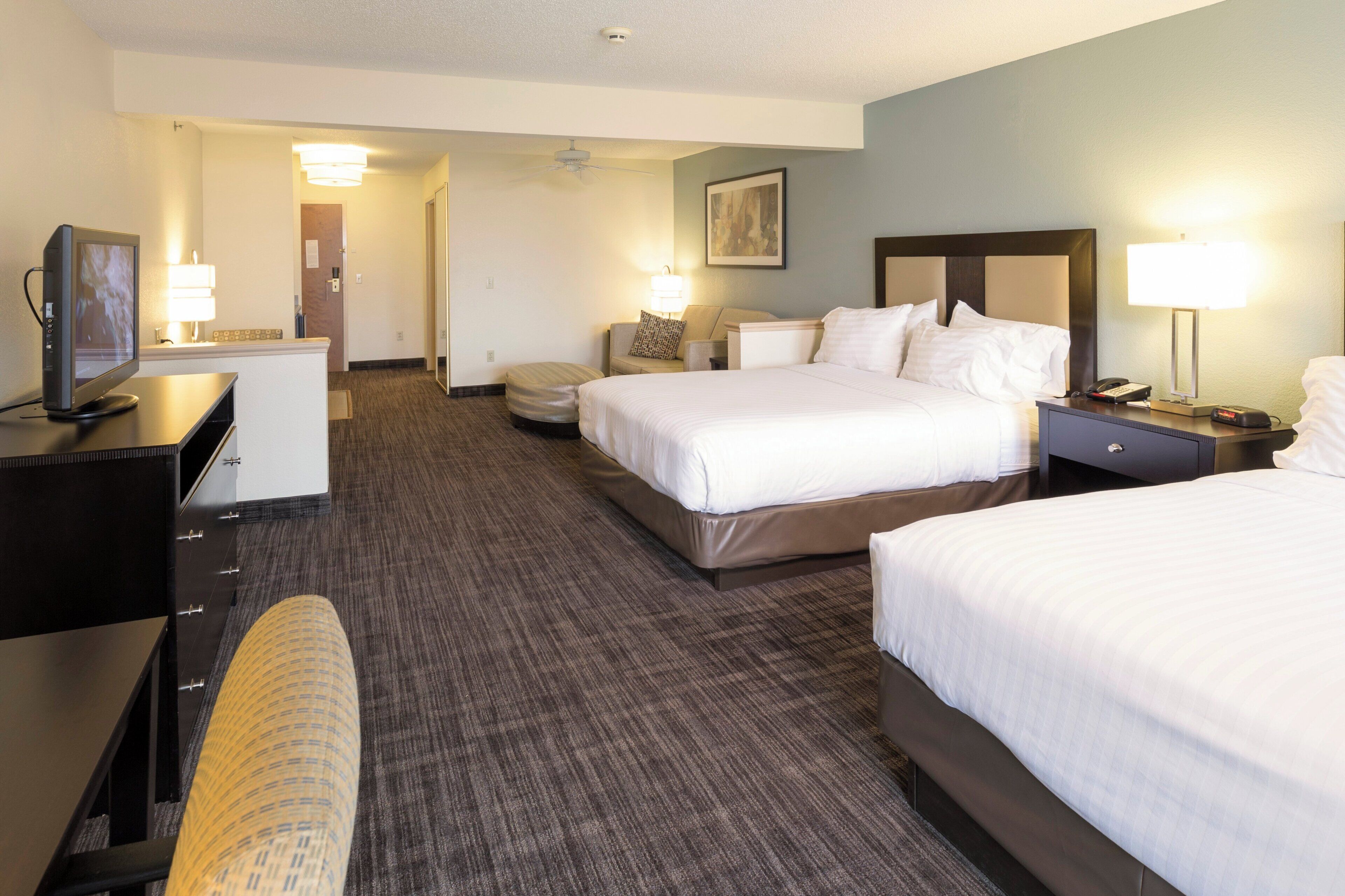 A hotel room with two beds, a desk with a computer, a chair, a bedside table with a lamp, and a wall-mounted TV.