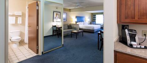 Habitación de hotel con baño, cama, escritorio, silla, cafetera y espejo.