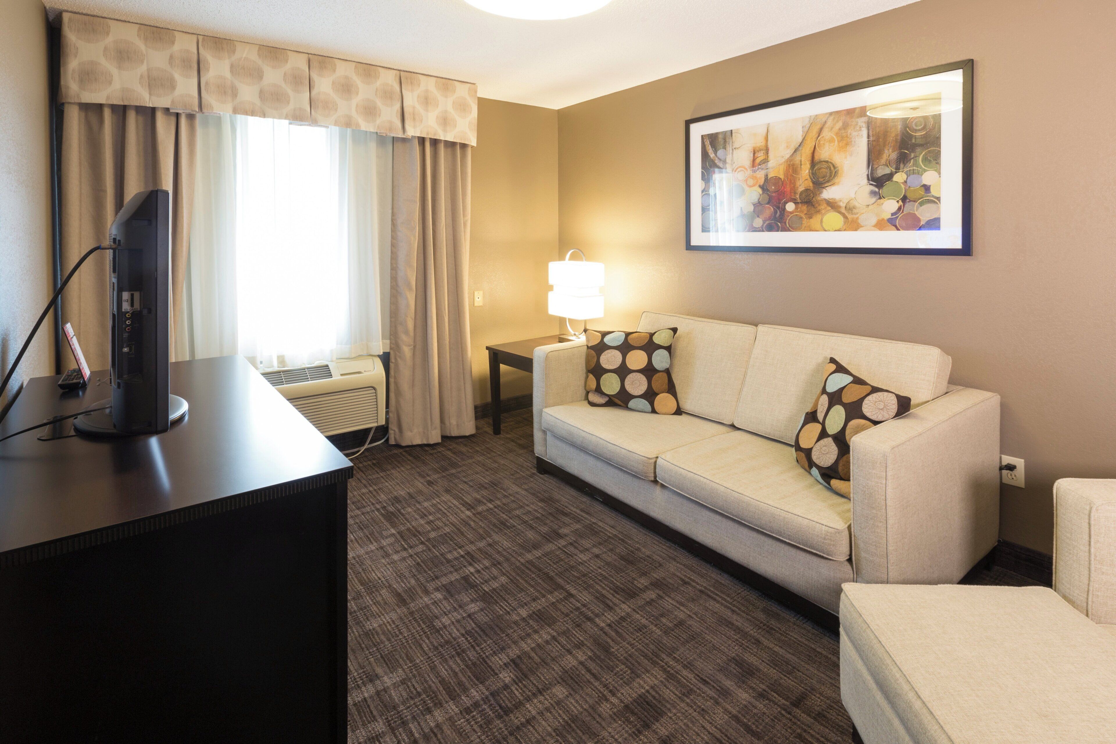 A hotel room with a flat-screen TV, a sofa with patterned cushions, a standing lamp, and a painting on the wall.