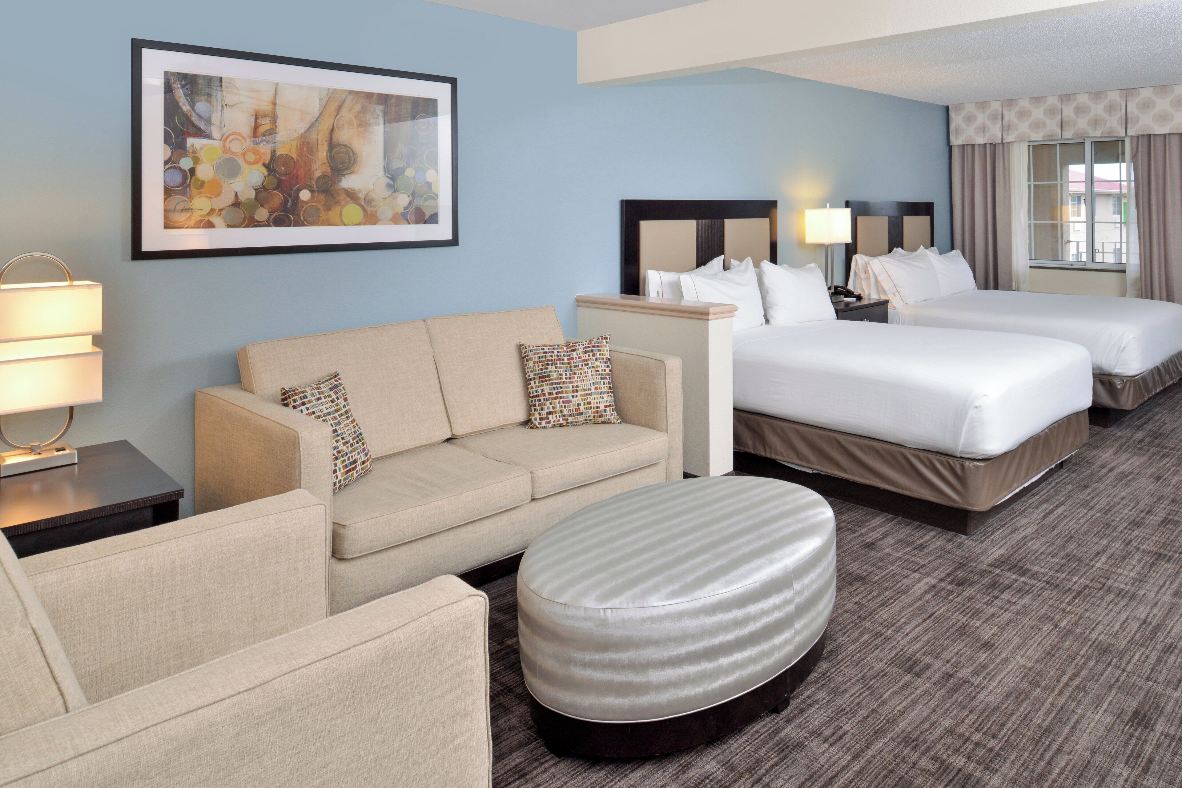 A hotel room with a sofa, two beds, a nightstand, a lamp, and a painting on the wall.