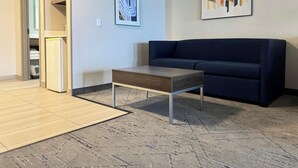 Un salon avec un canapé bleu foncé, une table basse en bois, un réfrigérateur et deux œuvres d’art encadrées accrochées au mur.