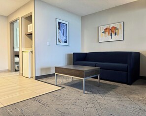 Un salon avec un canapé bleu foncé, une table basse en bois, un réfrigérateur et deux œuvres d’art encadrées accrochées au mur.