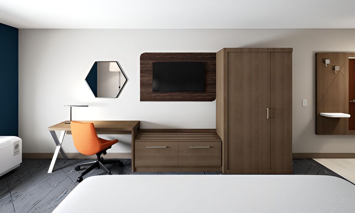 Quarto de hotel moderno com cama, escrivaninha com cadeira laranja, guarda-roupa, espelho e televisão fixada na parede.