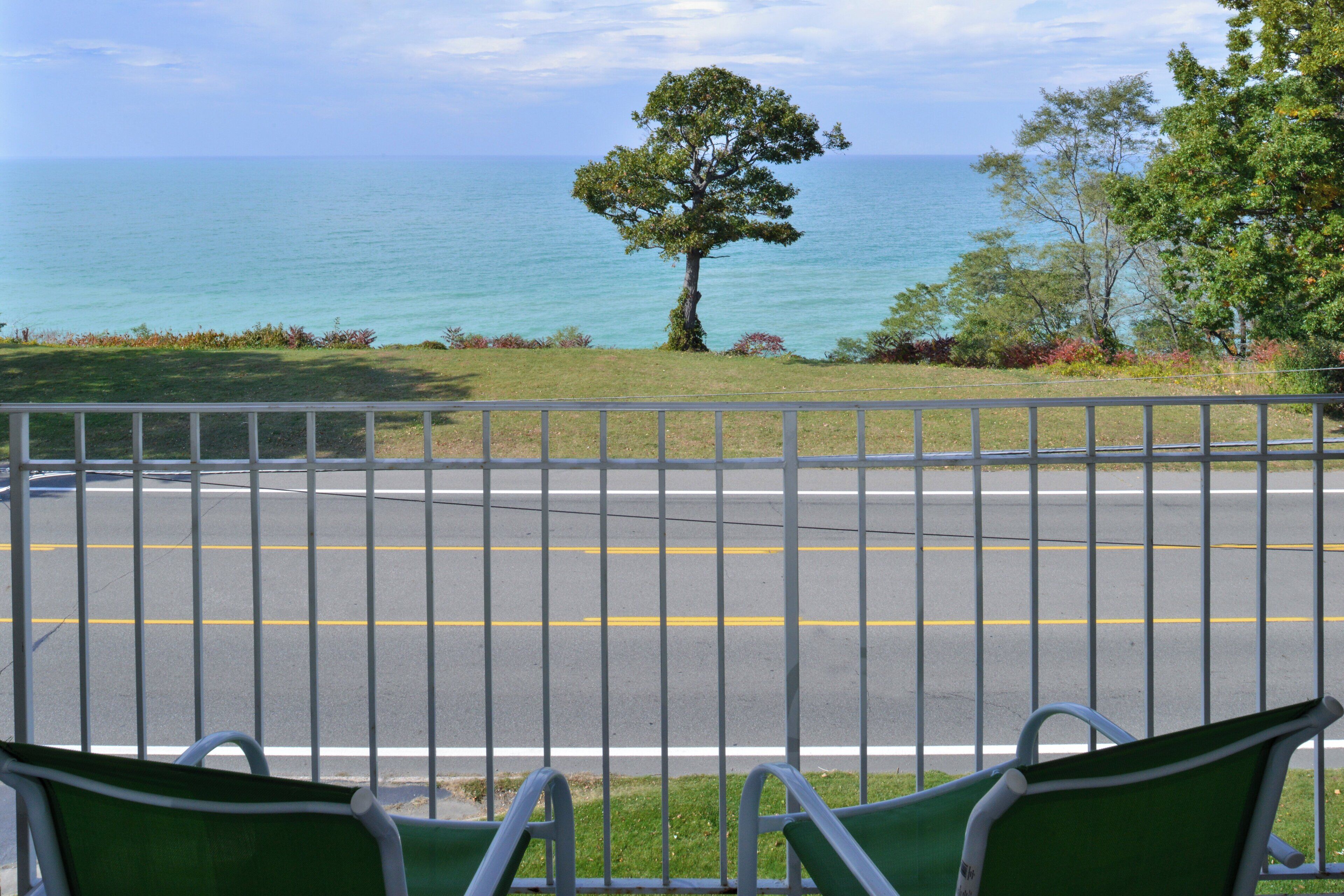 A view from a balcony with a metal railing, overlooking a road with yellow lines and a tree in the distance.