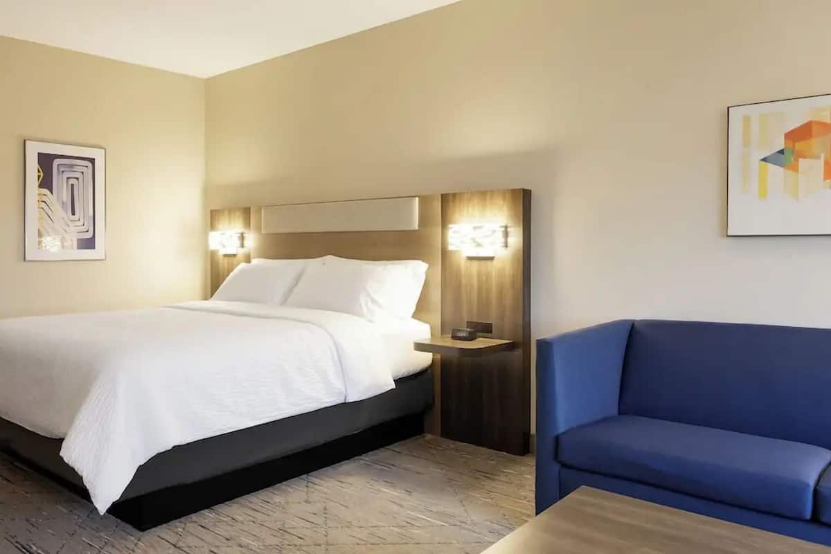 Habitación de hotel con una cama, un sofá azul, una mesita de noche y luces empotradas en la pared.