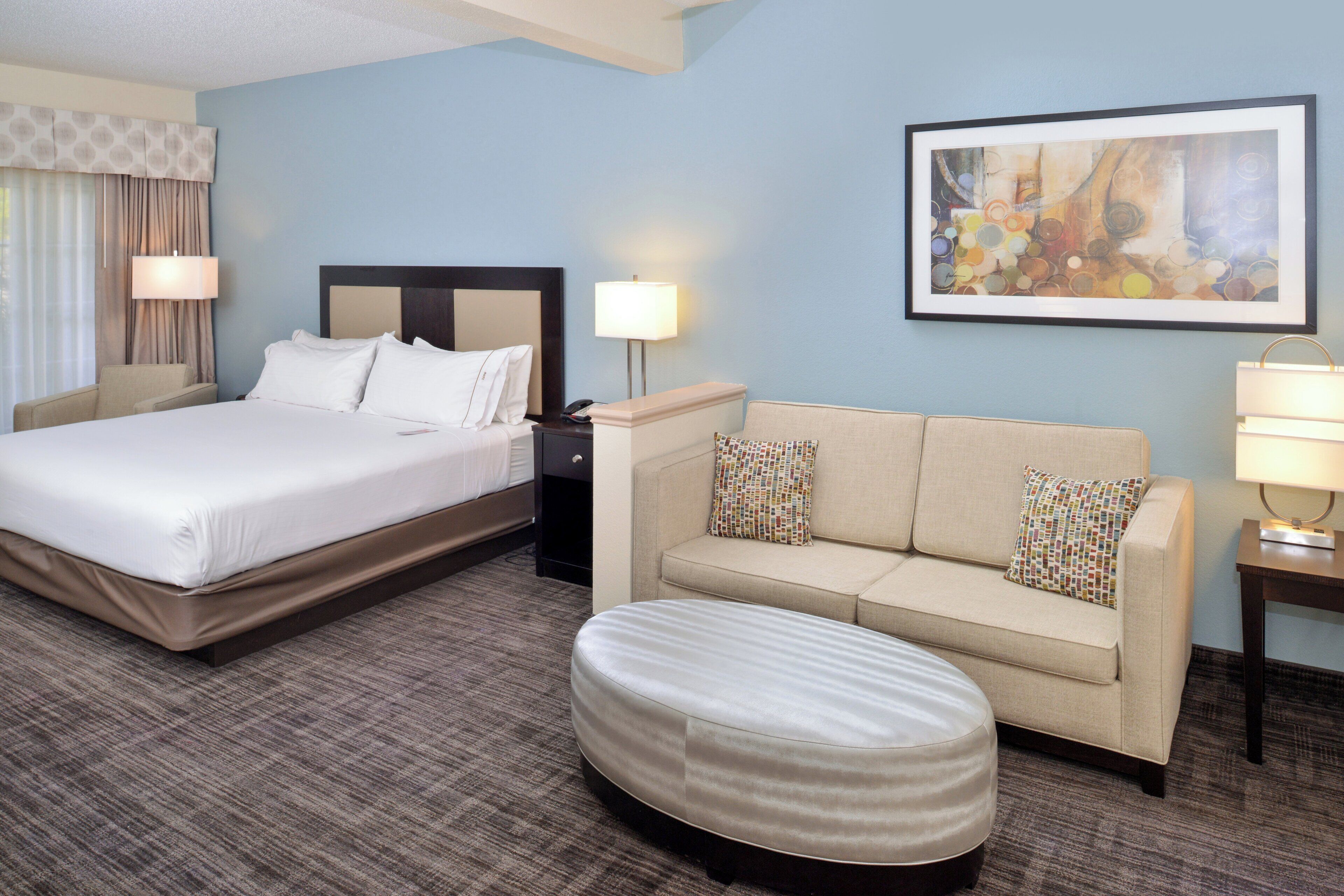 Habitación de hotel con cama, sofá, reposapiés, mesita de noche, lámpara y un cuadro en la pared.