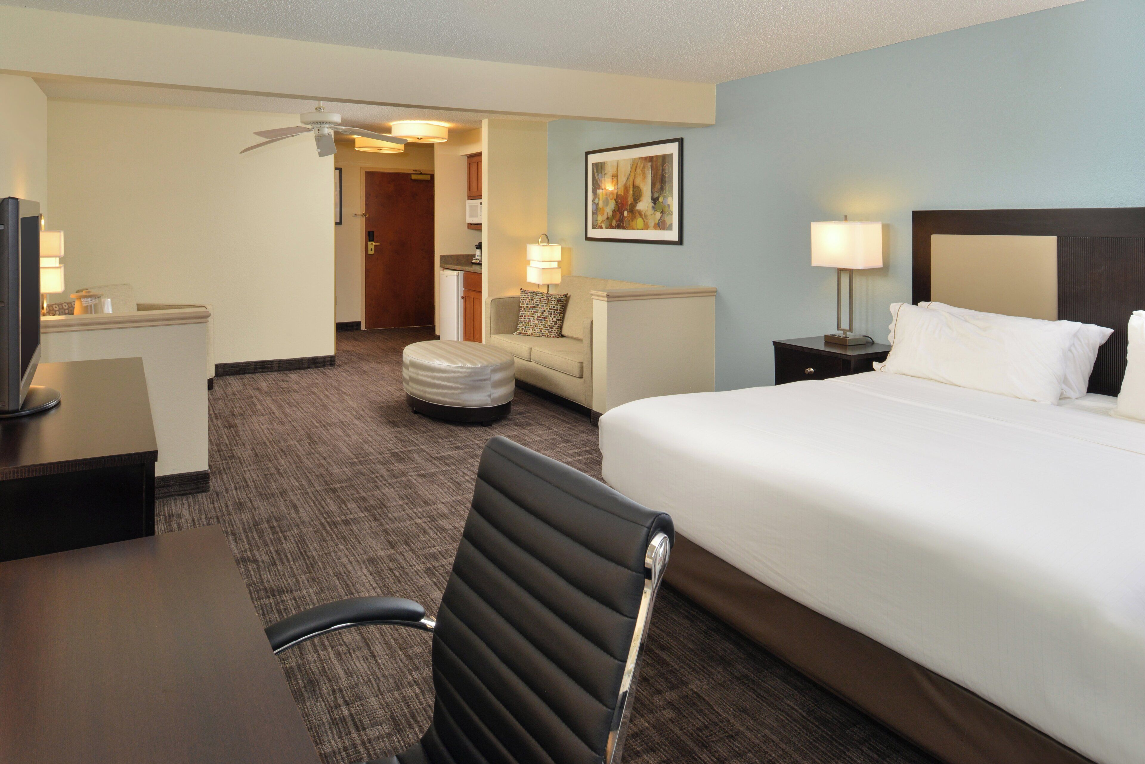 A hotel room with a bed, a desk, a chair, a television, a lamp, and a ceiling fan.