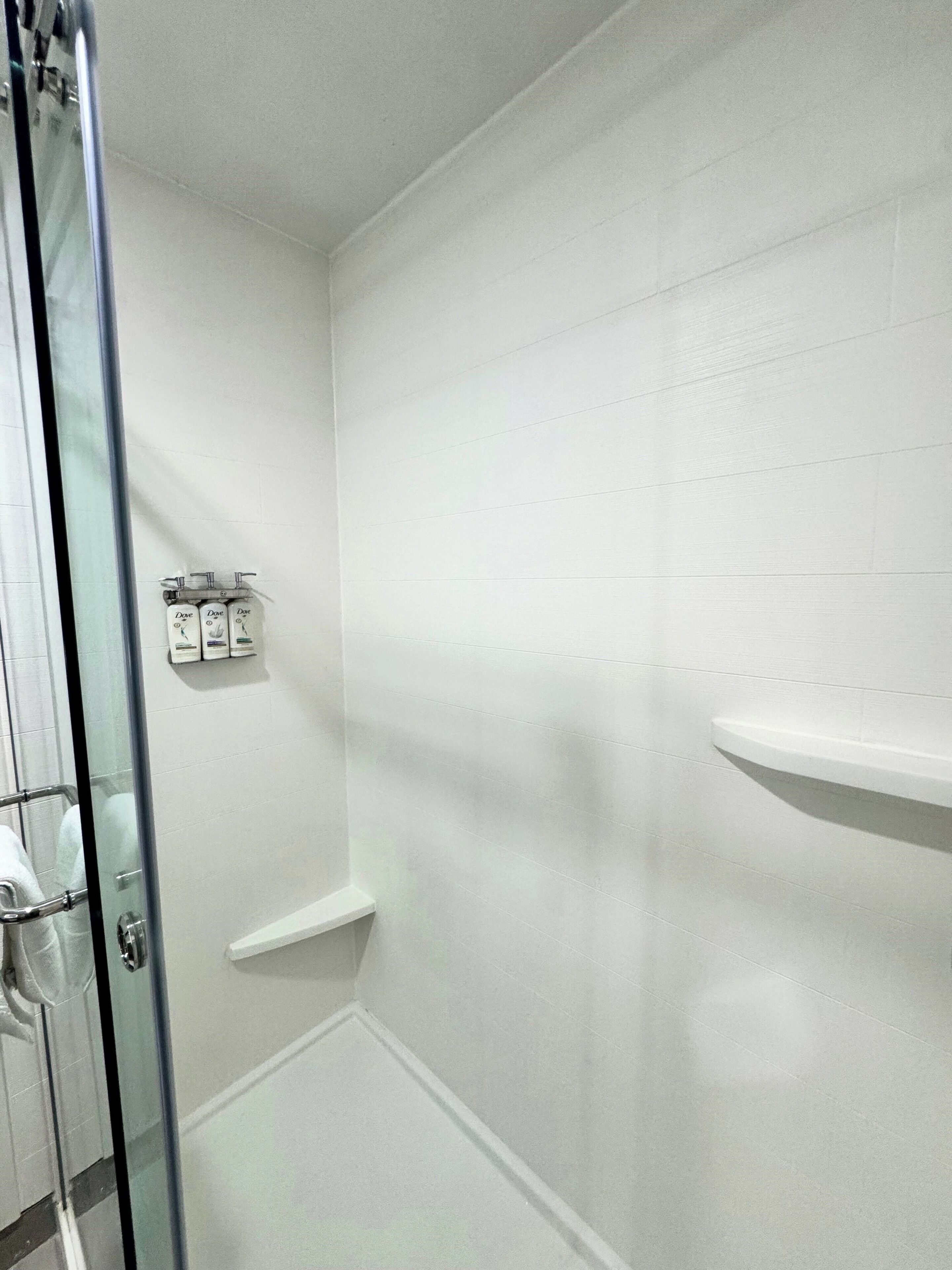 A bathroom with white tiled walls, a glass shower enclosure, and a shelf with toiletries.