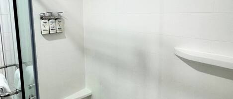 A bathroom with white tiled walls, a glass shower enclosure, and a shelf with toiletries.