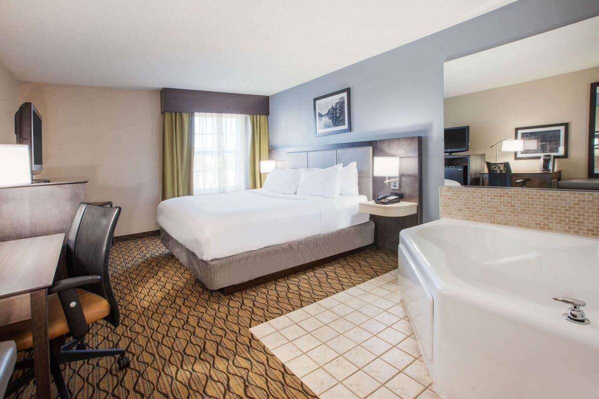 A hotel room with a bed, a desk, a chair, a desk lamp, a TV, a bathroom with a bathtub, and a window with curtains.