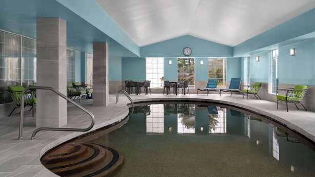 An indoor swimming pool with a tiled floor, a clock on the wall, and green chairs around the pool. - Hampton Inn Sturbridge (Sturbridge)