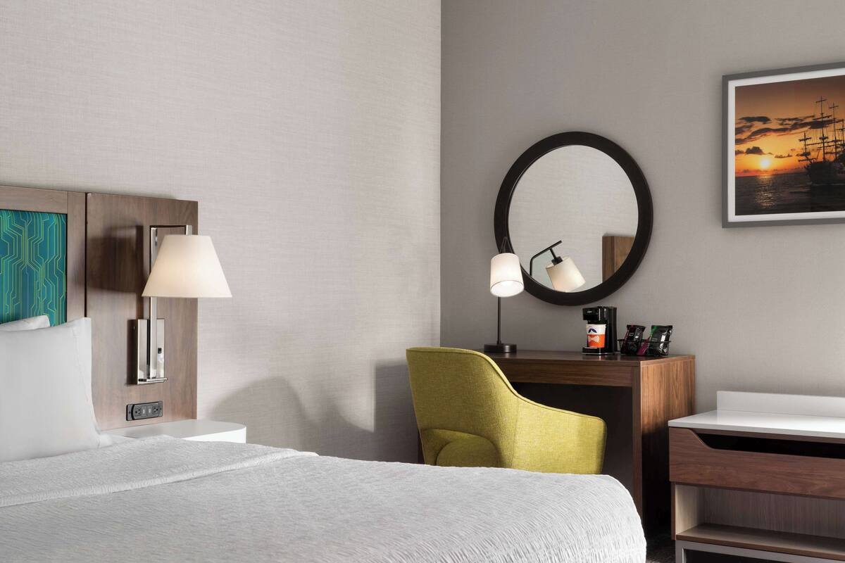 A hotel room with a bed, a desk, a chair, a lamp, a mirror, and a framed picture on the wall.