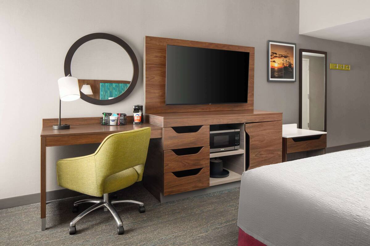 A hotel room with a wooden desk, a flat-screen TV, a yellow office chair, a bed with a white comforter, a microwave, a coffee maker, and a framed picture on the wall.