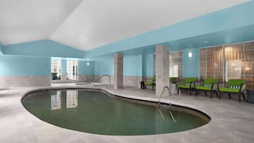 Una piscina con borde curvo, rodeada de un suelo con mosaicos y paredes pintadas en azul claro. Hay sillas verdes con cojines blancos dispuestas a lo largo de la pared.