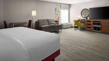 A hotel room with a bed, a sofa, a desk with a chair, a television, and a window with blinds.