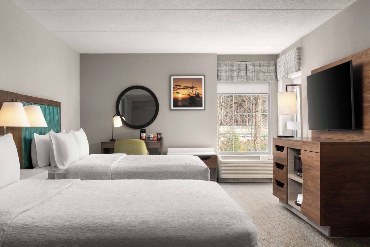 A hotel room with two beds, a TV, a desk with a lamp, a mirror, and a window with a view of trees.