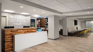 A reception area with a 'Welcome to the Patriot State' sign, a reception desk, a minibar, and a seating area with yellow chairs. - Hampton Inn Sturbridge (Sturbridge)