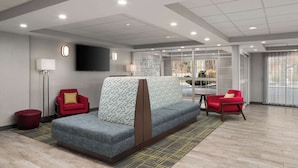 A modern hotel lobby with a grey sofa, red armchairs, a flat-screen TV, and a large window offering a view of the outdoors. - Hampton Inn Sturbridge (Sturbridge)