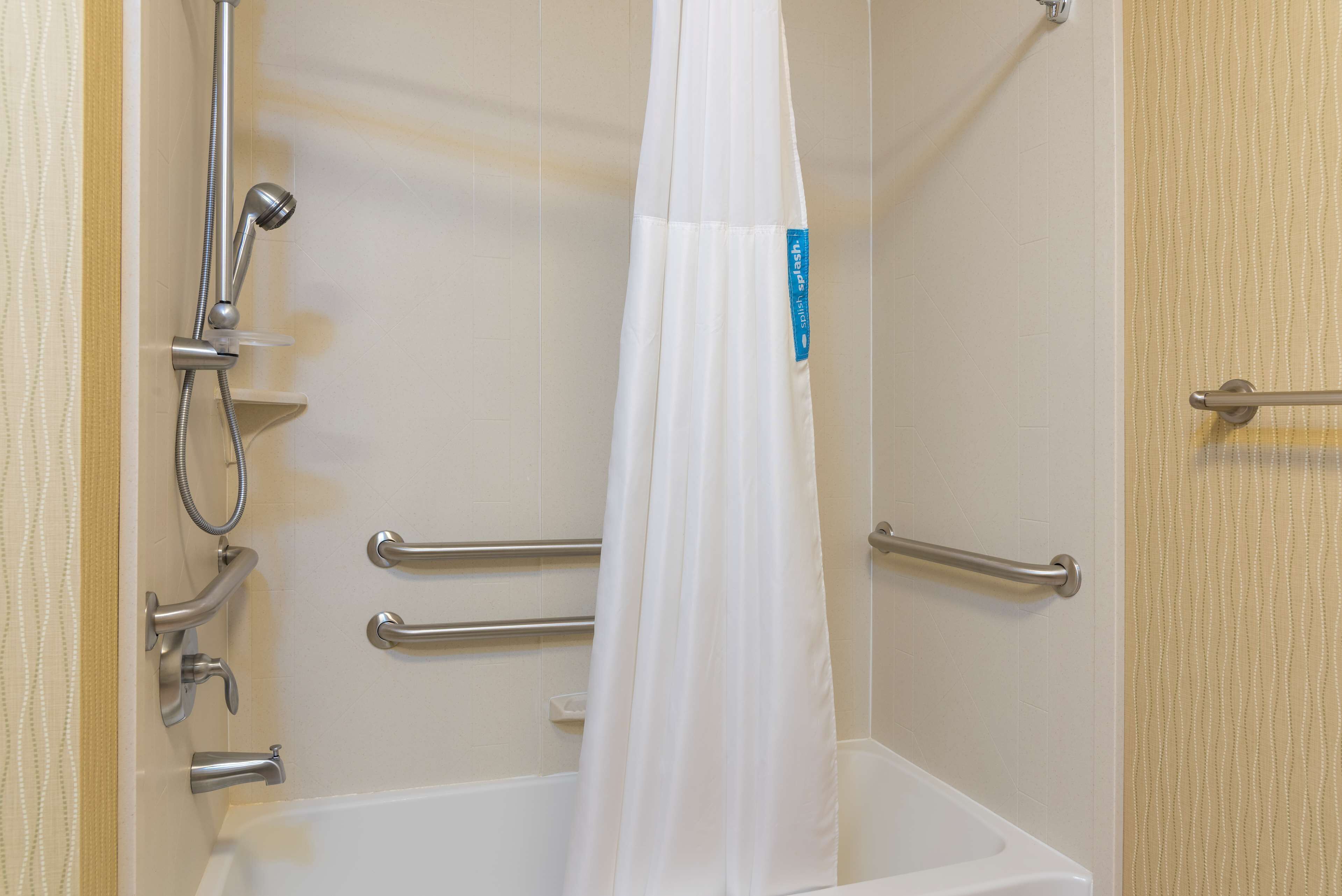 Un espace douche doté d’un rideau blanc, de barres d’appui et d’un pommeau de douche amovible.