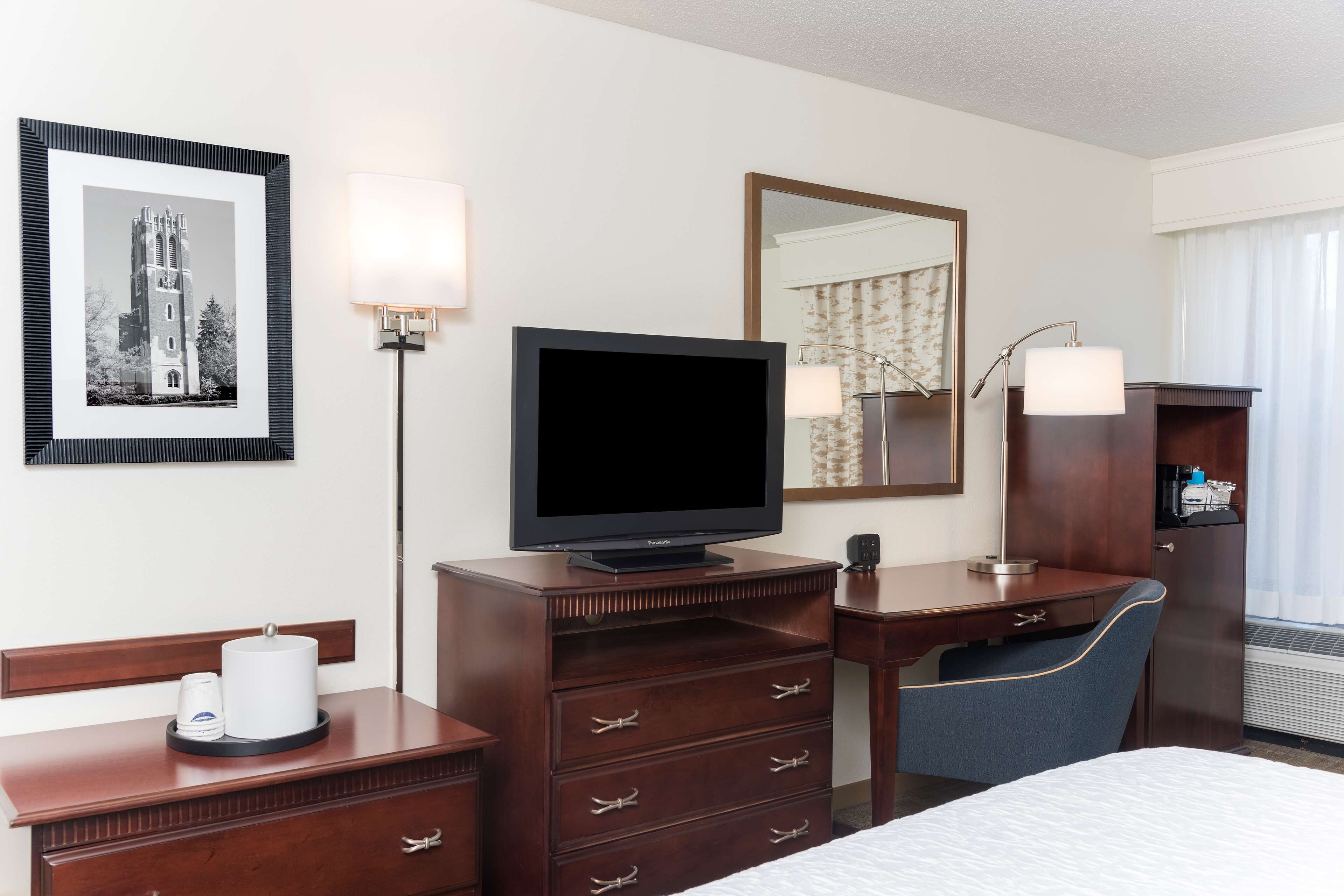 A hotel room with a bed, a desk with a chair, a television, a dresser, a mirror, and a framed picture on the wall.