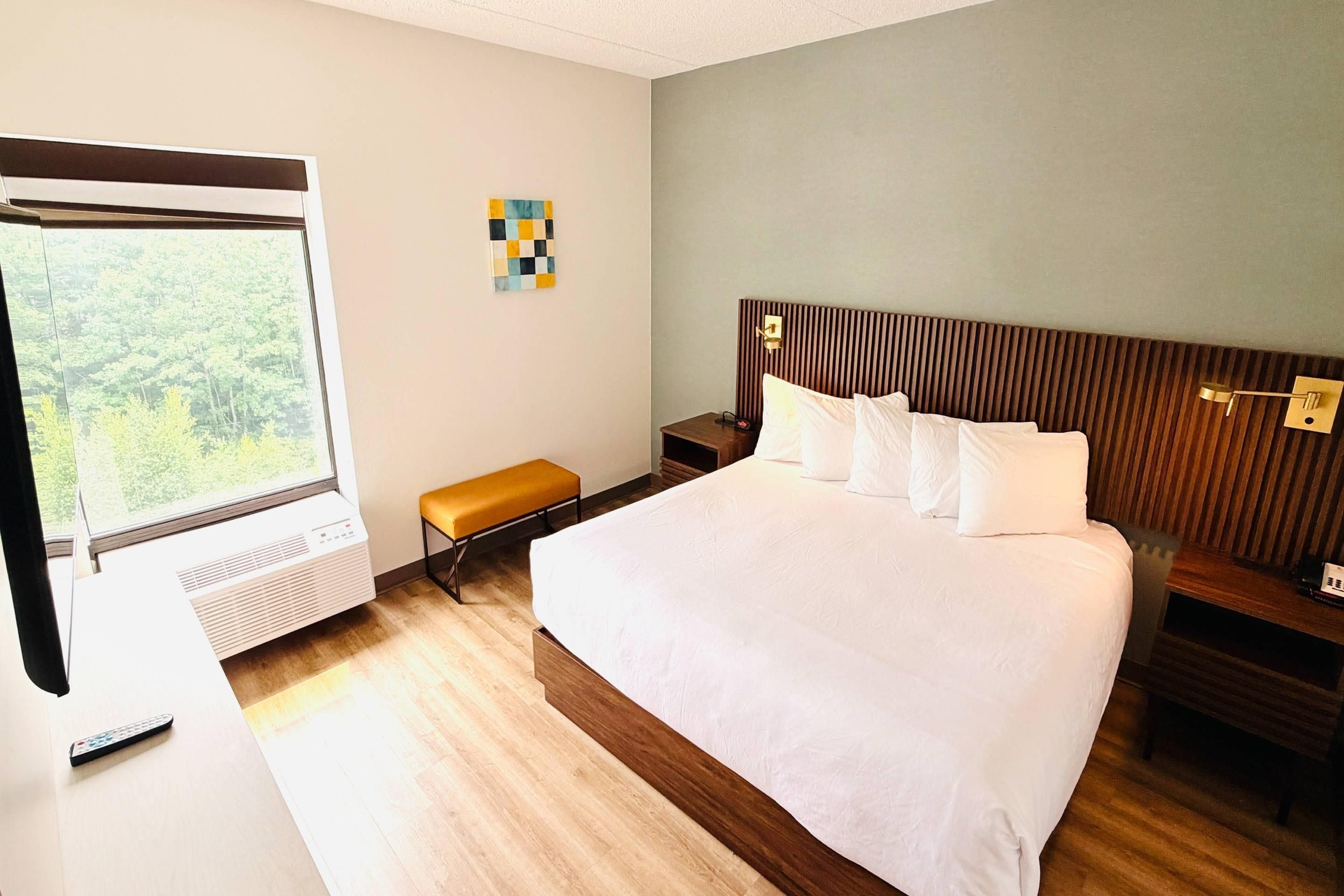 A hotel room with a large bed, a bedside table, a bench, a wall-mounted air conditioner, and a window with a view of trees.