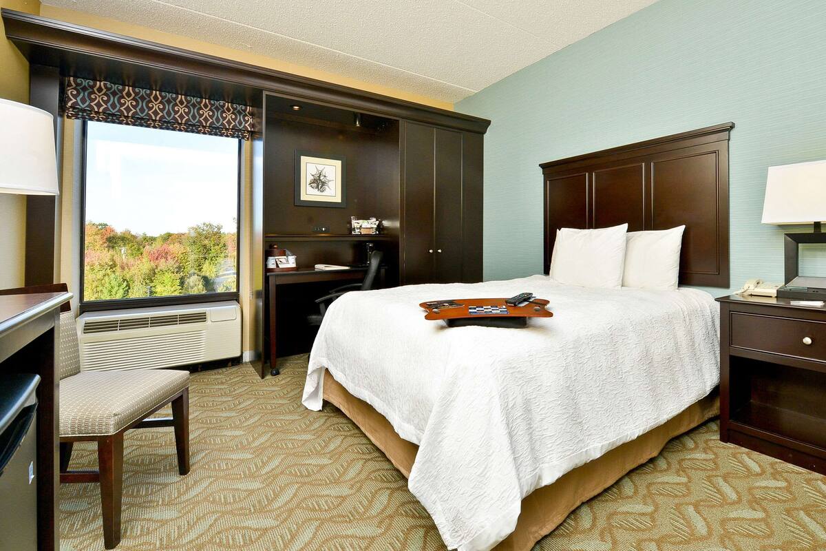 A hotel room with a large bed, a desk, a chair, and a view of trees outside the window.