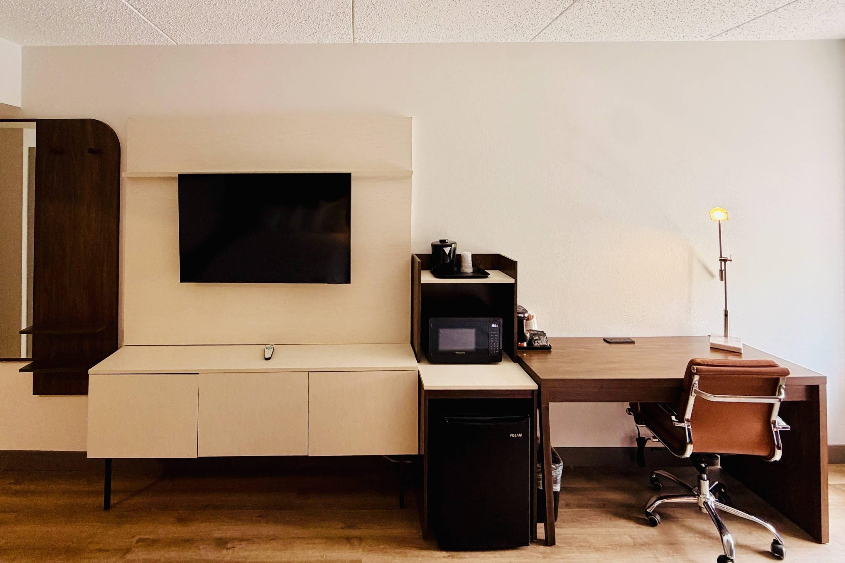 A modern office space with a flat-screen TV mounted on the wall, a desk with a computer, a microwave, and a chair.