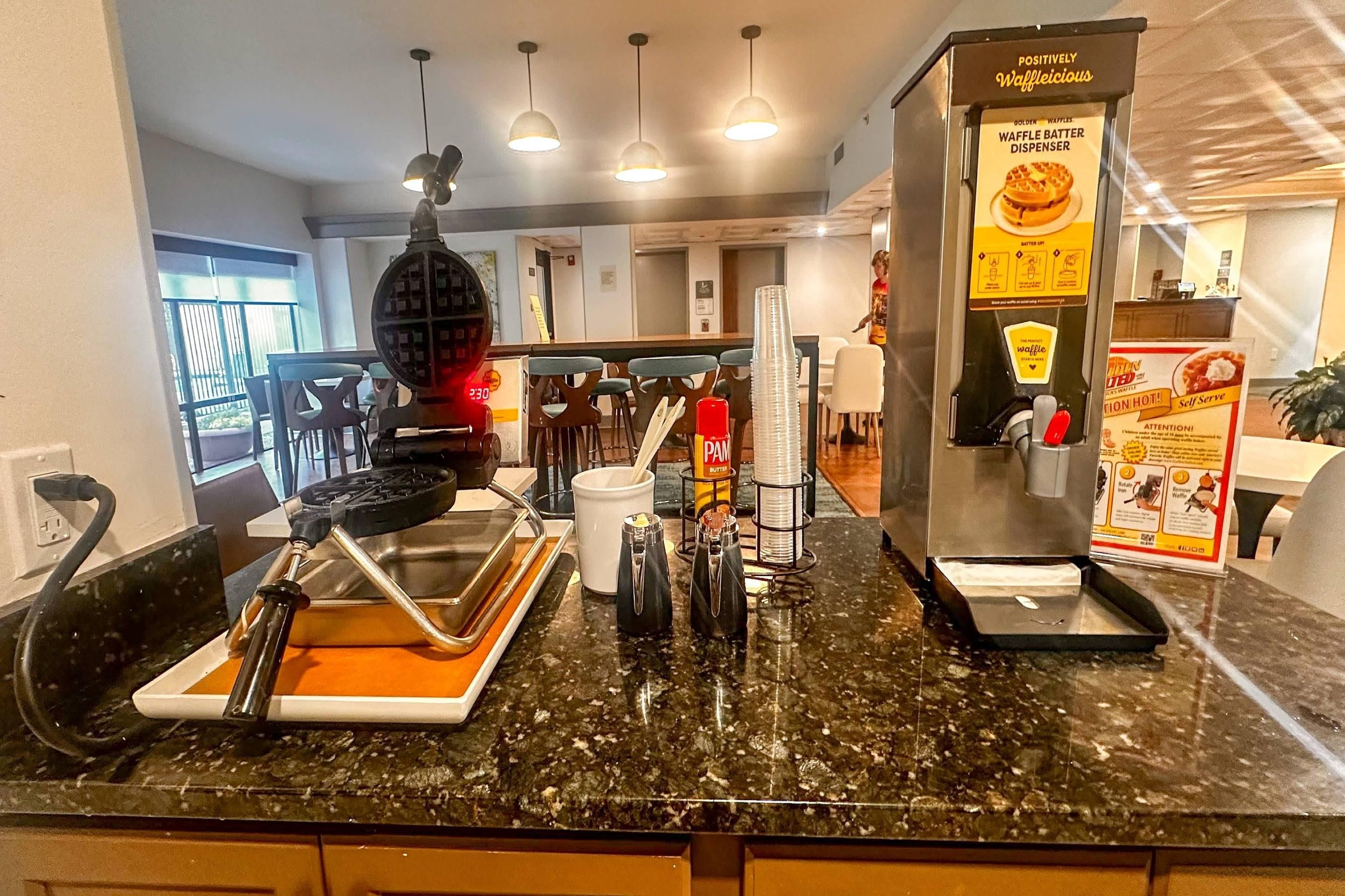 Una zona de cocina con dispensador de masa para gofres, gofarrera y cafetera.
