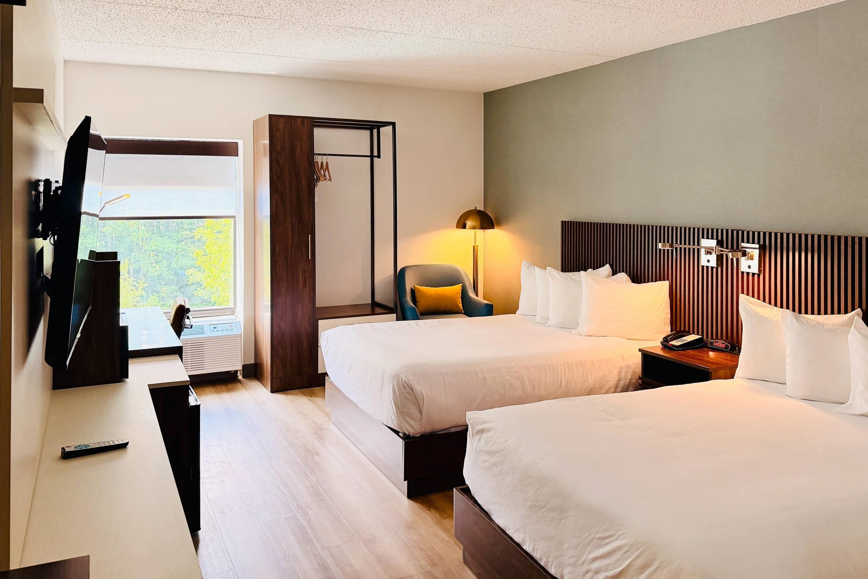 Une chambre d’hôtel avec deux lits, un bureau avec une chaise, une télévision et une fenêtre donnant sur des arbres.