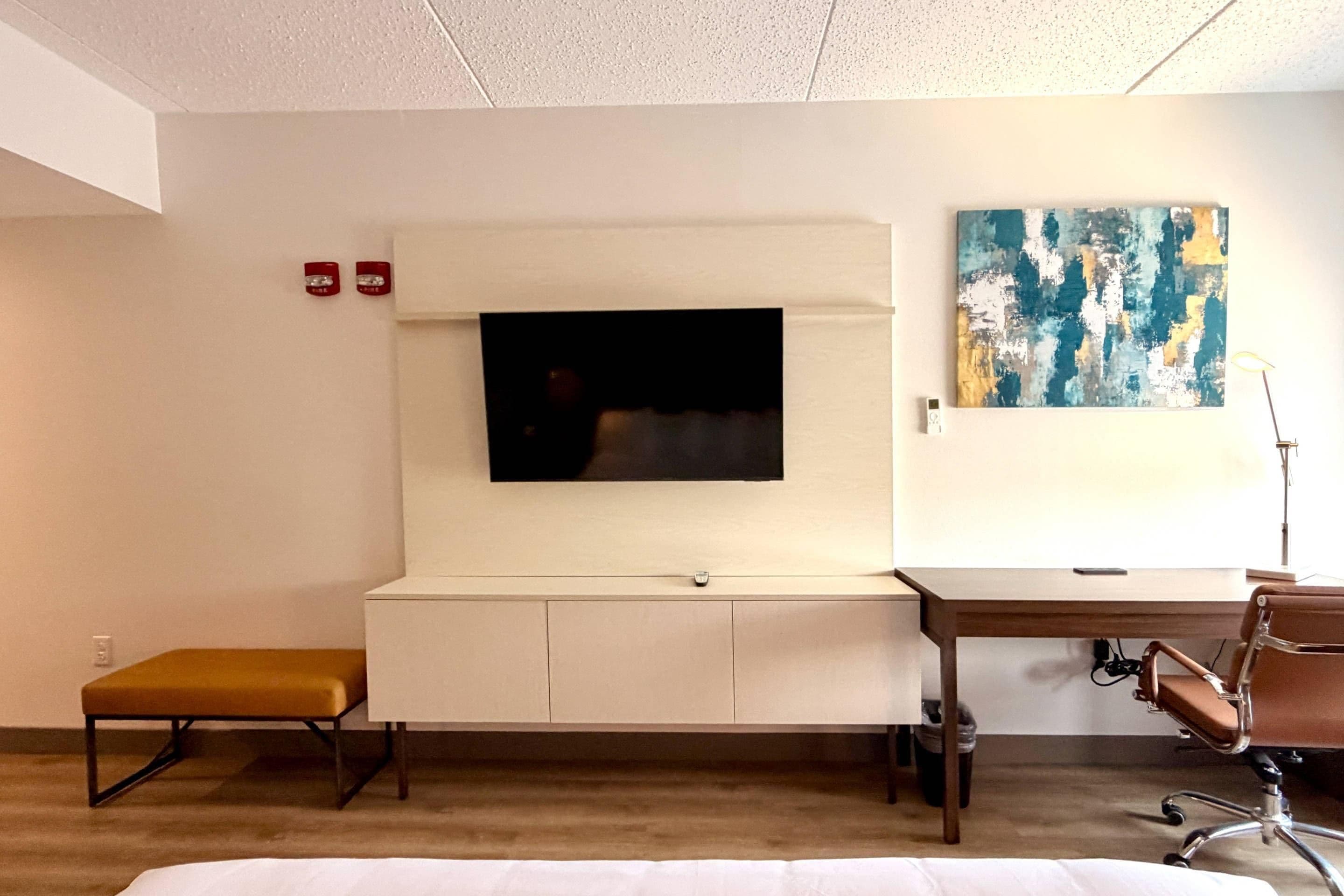 A hotel room with a flat-screen TV mounted on a white entertainment unit, a desk with a chair, and a small bench.