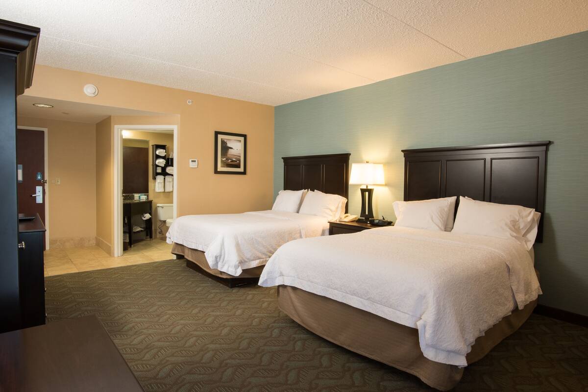 A hotel room with two beds, a nightstand with a lamp, a bathroom visible through an open door, and a small desk.