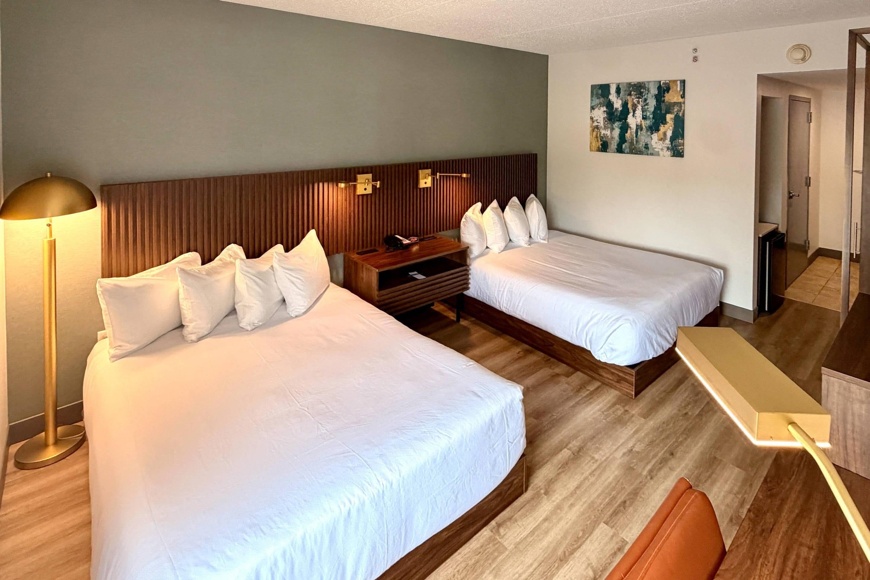 Ein Hotelzimmer mit zwei Betten, einem Schreibtisch, einer Lampe und einem Gemälde an der Wand.