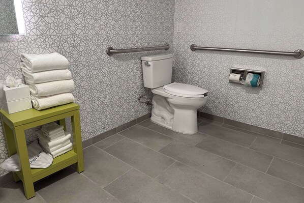 A bathroom with a toilet, a stack of white towels, a hand dryer, and a hand sanitizer dispenser.