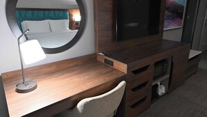 A hotel room with a wooden desk, a round mirror, a flat-screen TV, a bed with white linens, and a painting on the wall.