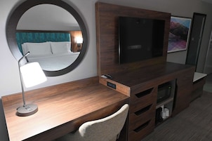 A hotel room with a wooden desk, a round mirror, a flat-screen TV, a bed with white linens, and a painting on the wall.