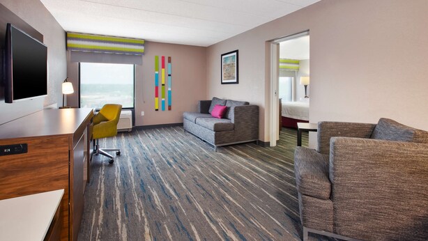 A hotel room with a large TV, a desk, a sofa, and a chair. There is a window with a view of the city. - Hampton Inn College Park (College Park)
