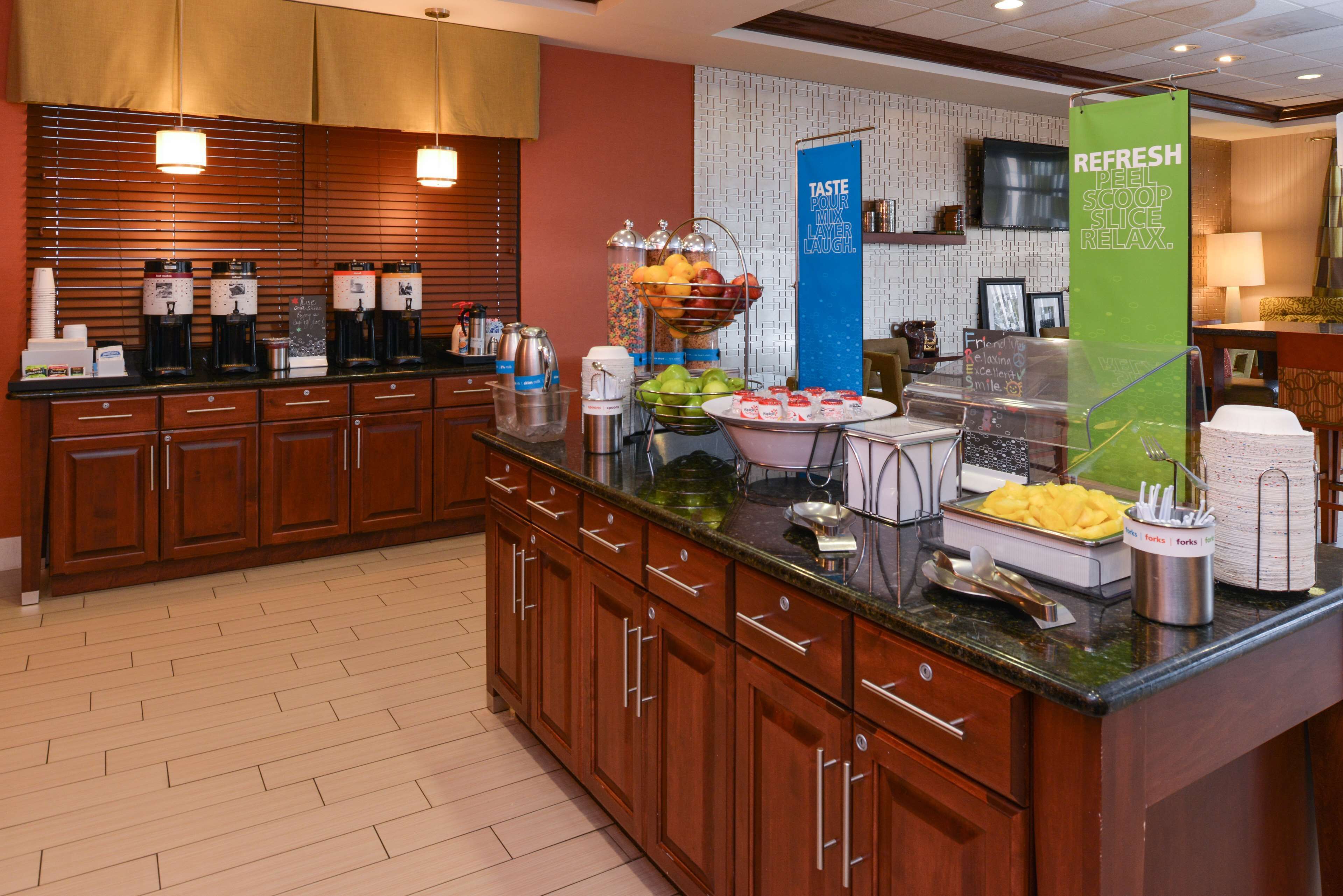 A hotel kitchen with a buffet setup, including coffee makers, fruit bowls, and a 'TASTE POUR MIX LAUGH' sign.
