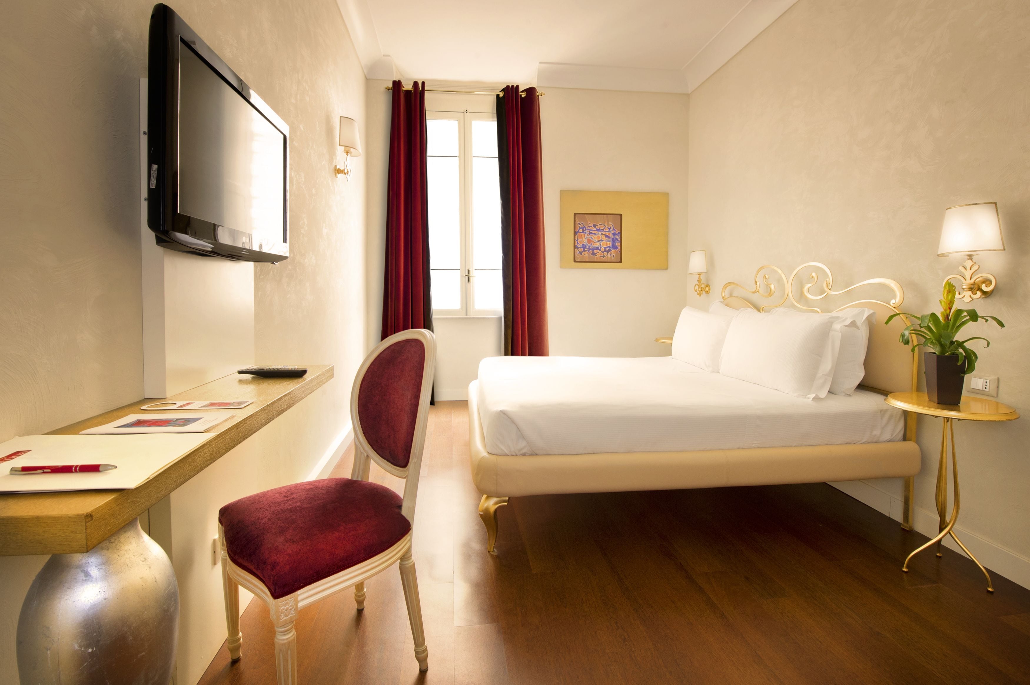 A hotel room with a bed, a desk with a chair, a television, and a small table with a lamp and a plant.