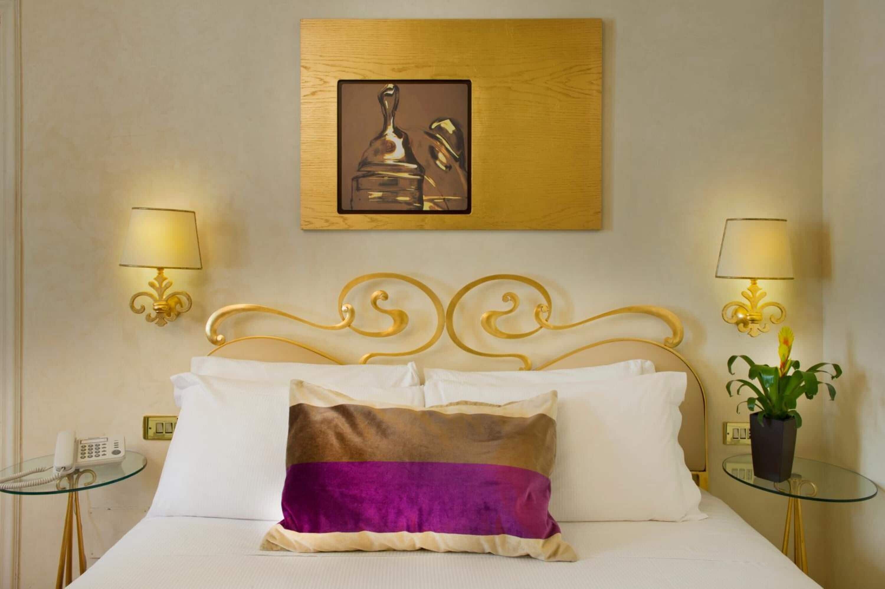 A hotel room with a bed, two bedside lamps, a wall-mounted painting, a telephone, and a small table with a plant.