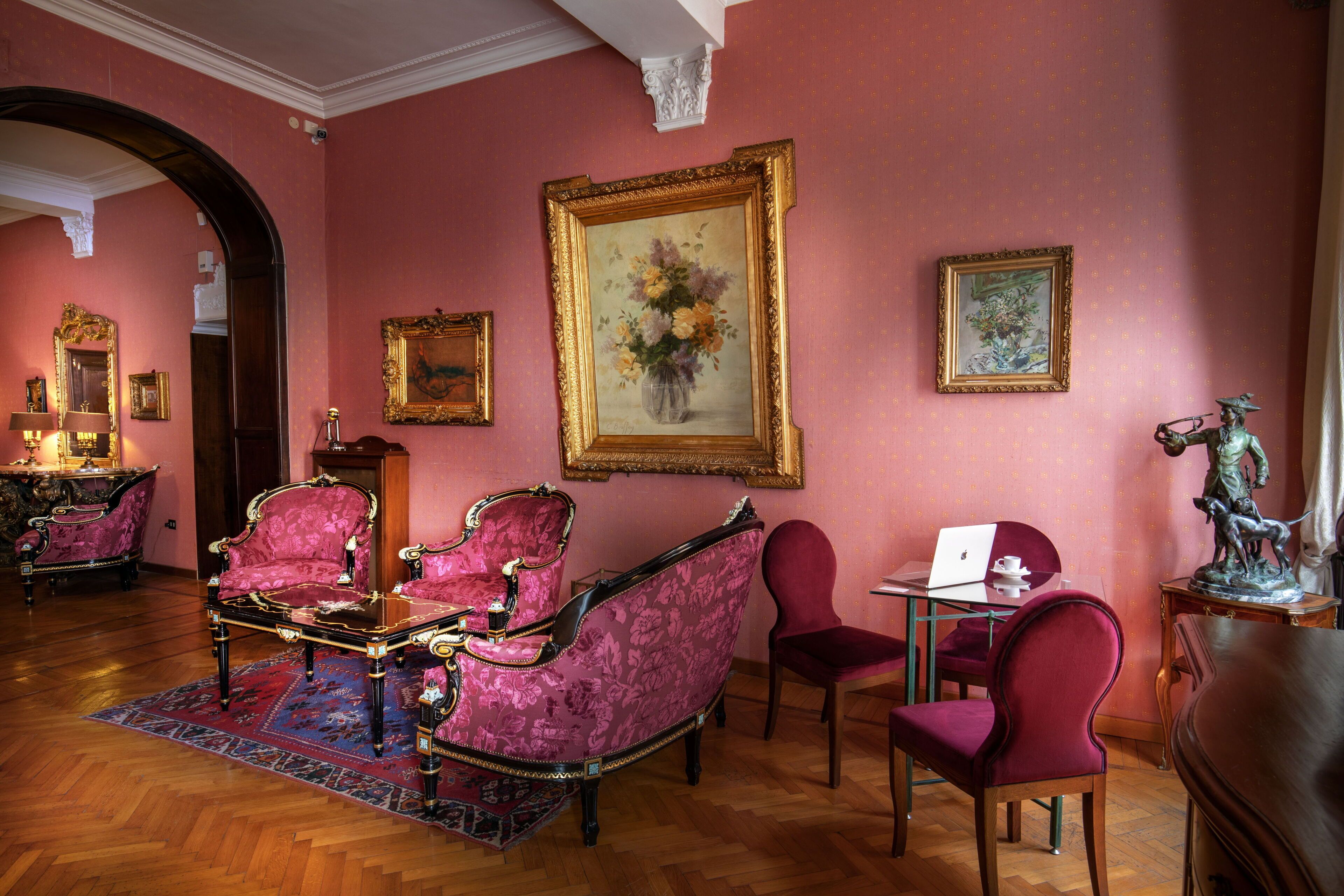 Una stanza con pavimento in legno, pareti rosa e un grande quadro di fiori. Si notano due sedie, un tavolo e una statua.