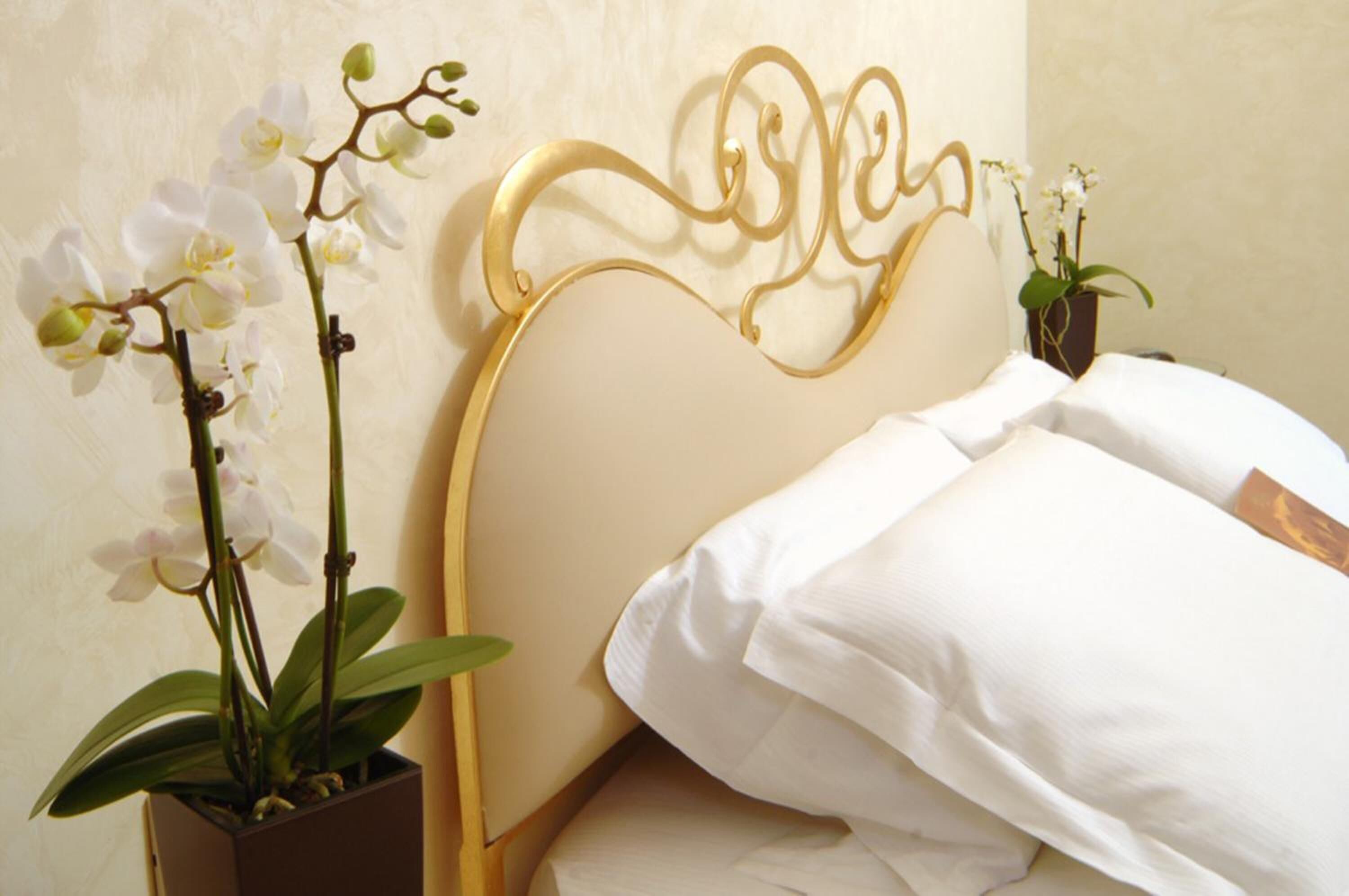 A neatly made bed with white linens, a gold ornate headboard, and two potted orchids on either side.