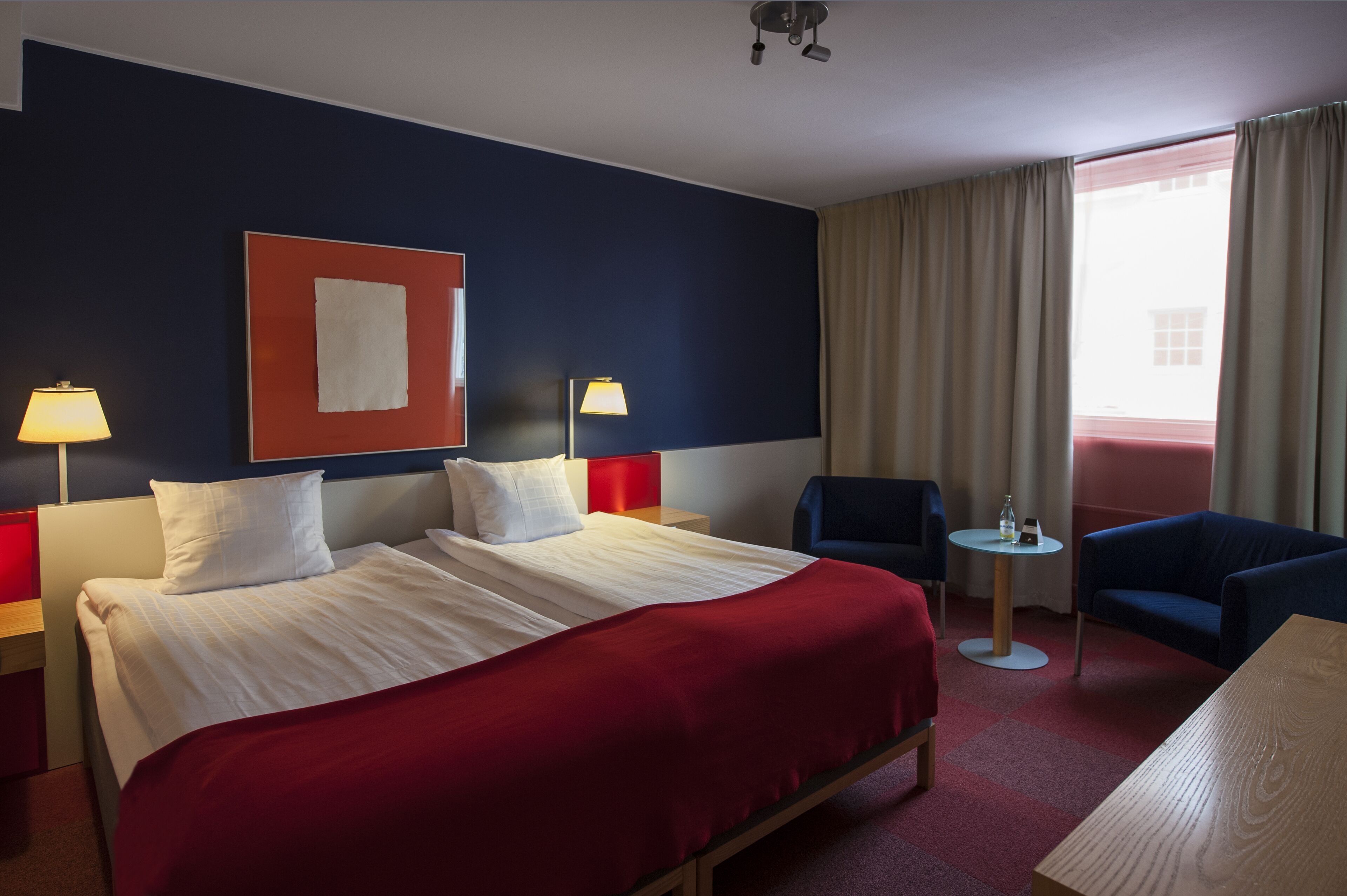 Habitación de hotel con una cama grande, dos mesitas de noche con lámparas, una pequeña mesa redonda, un sillón azul y vistas al exterior a través de una ventana con cortinas.