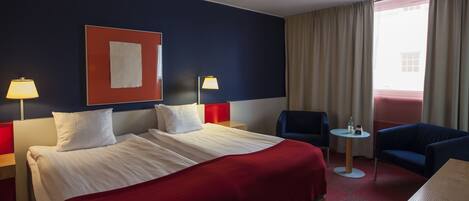 Quarto de hotel com uma cama grande, duas mesas de cabeceira com luminárias, uma pequena mesa redonda, uma poltrona azul e vista para o exterior através de uma janela com cortinas.