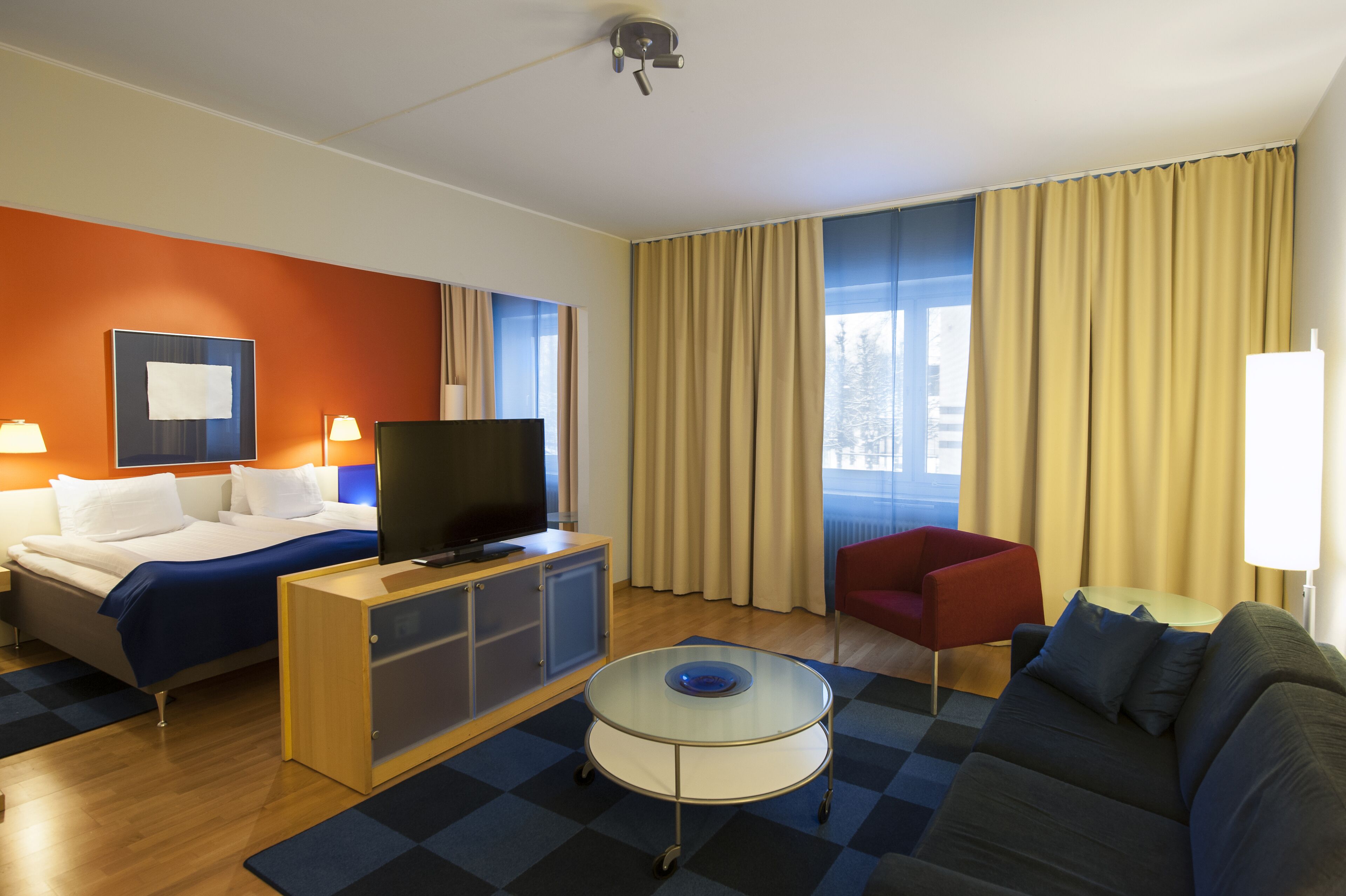 Una habitación de hotel moderna con una cama, un televisor, un sillón rojo, un sofá y una mesa de centro de vidrio.