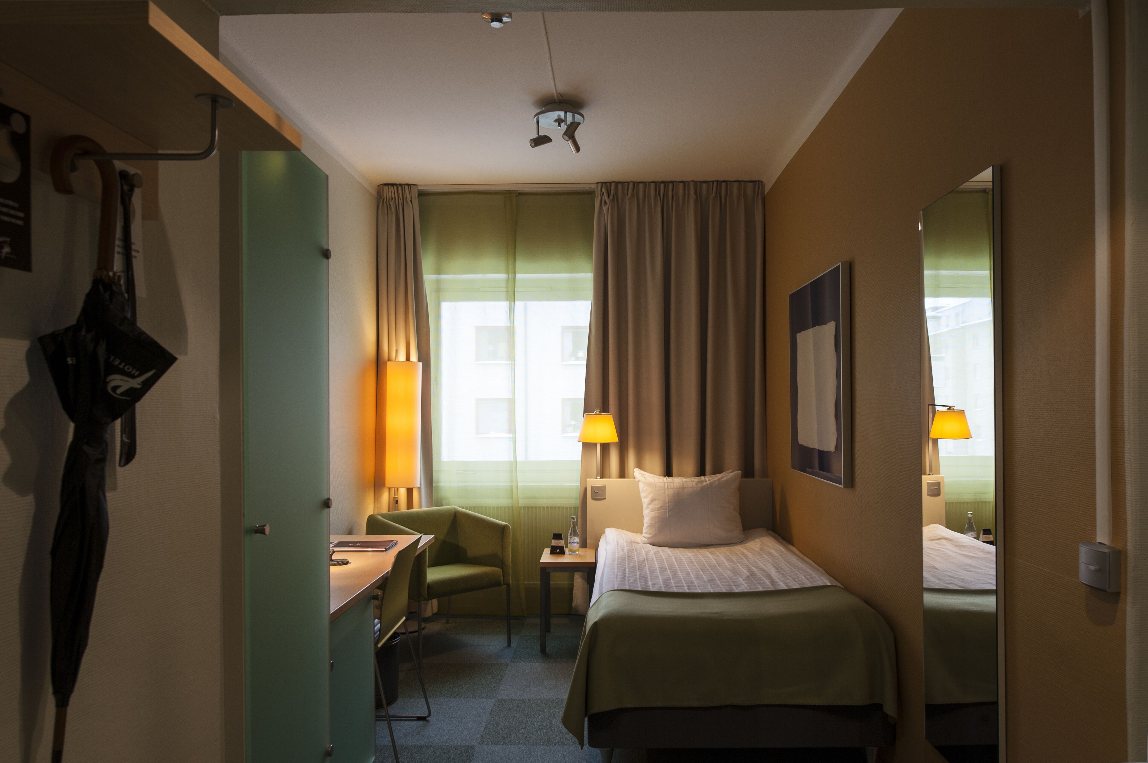 Une chambre d’hôtel avec un lit, un bureau, une chaise, une lampe et une fenêtre avec des rideaux.